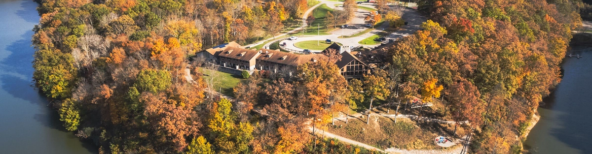 Aerial view of a large lodge surrounded by colorful autumn trees, located on a forested peninsula jutting into a calm lake, with parking lots and winding roads visible.