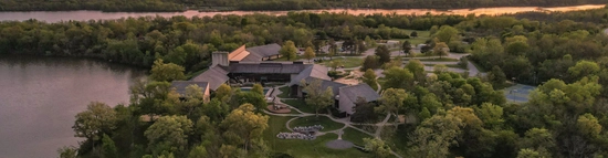 Aerial view of a large lodge surrounded by dense green trees, located by a calm lake at sunset, with winding paths and outdoor spaces visible around the building.