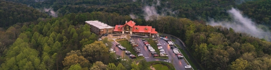 Aerial view of a large lodge with a red roof surrounded by dense green forest; numerous cars are parked in front, and low clouds or mist linger among the trees.
