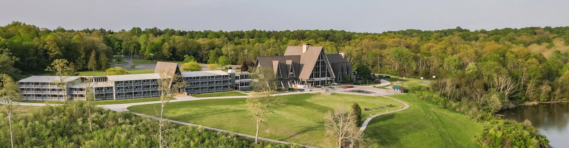 Aerial view of a large lodge with a modern building and triangular-roofed structure, surrounded by green trees, lawns, and a nearby lake under a clear sky.