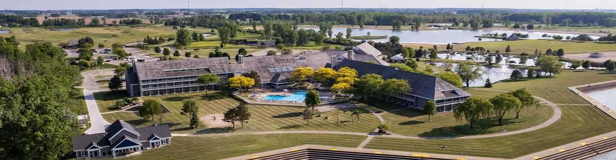 Aerial view of a large resort complex with a central outdoor pool, surrounded by green lawns, trees, ponds, and winding paths, set in a rural landscape with lakes and wooded areas in the distance.