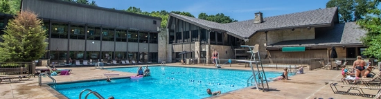 Outdoor swimming pool with people enjoying the water, surrounded by lounge chairs. Modern lodge-style buildings with large windows and sloped roofs are in the background, with trees and blue sky visible.