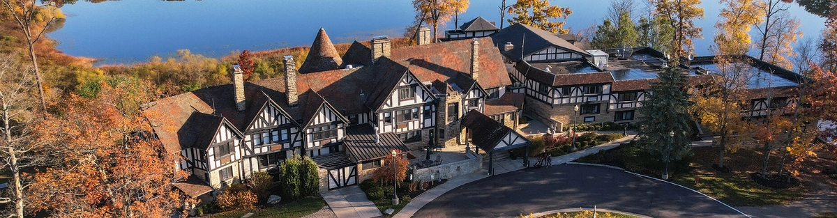 A large Tudor-style mansion with steep gabled roofs sits among autumn trees by a calm lake, viewed from above on a sunny day. A curved driveway leads to the building’s entrance.