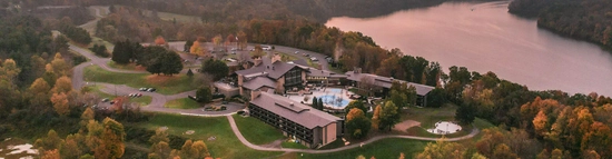 Aerial view of a large lakeside resort surrounded by autumn trees, with a pool at the center, winding roads, and a calm lake in the background under a pinkish sky.