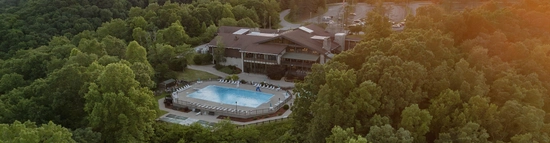 Aerial view of a large lodge surrounded by dense green forest, featuring a rectangular outdoor swimming pool with lounge chairs arranged around it. Sunlight streams in from the right side of the image.