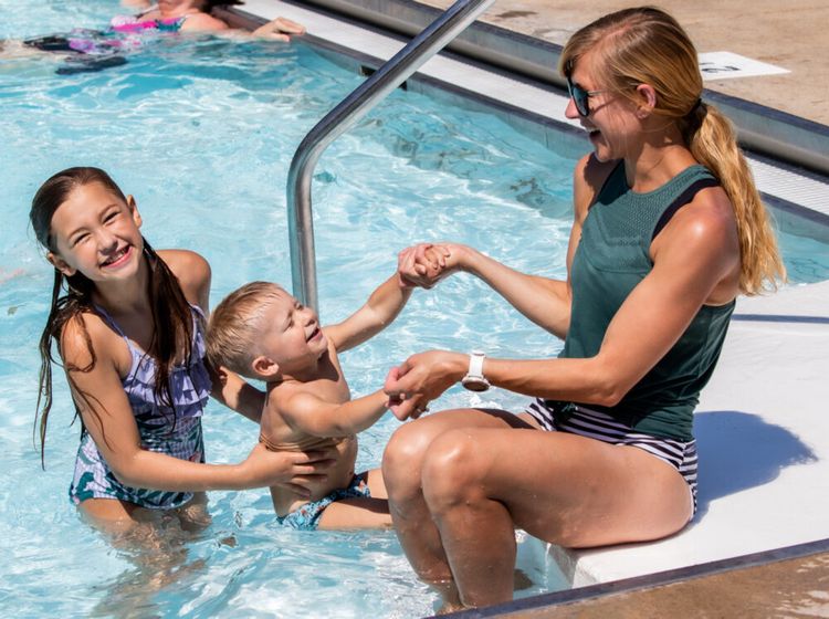 A woman sits on the edge of a pool, smiling and holding hands with a young boy in swim trunks, while a girl in a swimsuit stands in the water beside them, all appearing happy and playful.