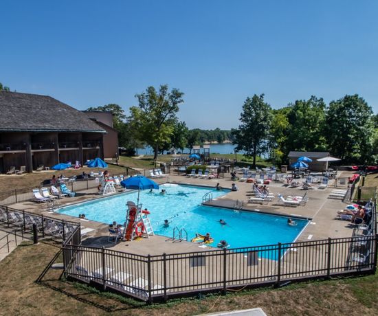 A rectangular outdoor swimming pool with people swimming and relaxing on lounge chairs, surrounded by two buildings. Trees and a lake are visible in the background under a clear blue sky.