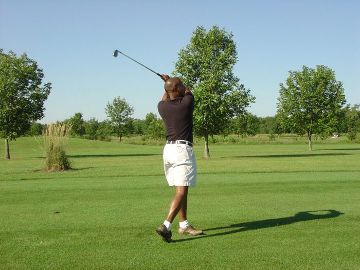A man wearing a black shirt, white shorts, and brown shoes swings a golf club on a grassy golf course with scattered trees under a clear blue sky.