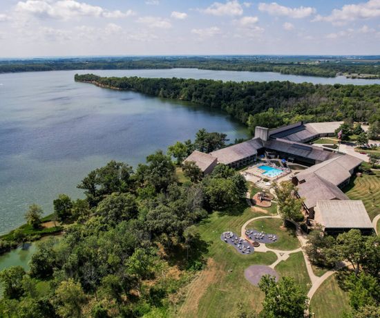 Aerial view of a lakeside resort surrounded by trees, with outdoor swimming pools, walking paths, and buildings extending along the shoreline under a partly cloudy sky.