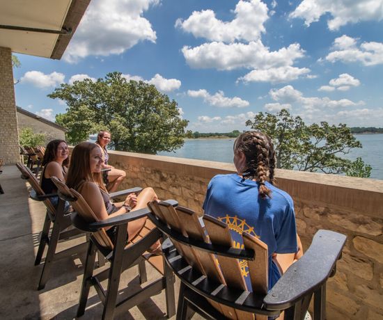 Four people sit on wooden chairs, relaxing and talking on a stone balcony overlooking a scenic lake with trees and a blue sky filled with fluffy clouds.