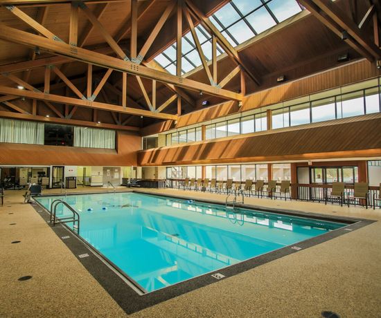 A spacious indoor swimming pool with clear blue water, surrounded by chairs and tables, beneath high wooden ceilings with large windows letting in natural light.