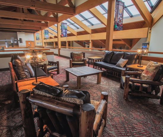 A cozy loft seating area with rustic wooden furniture, black leather cushions, lamps on end tables, and large windows in a vaulted ceiling letting in natural light. Carpeted floor with a patterned design.