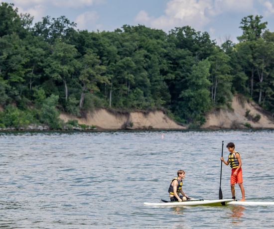 Two people wearing life jackets are on a paddleboard in a lake; one is sitting while the other stands and paddles. Green trees and sandy cliffs are visible in the background under a partly cloudy sky.
