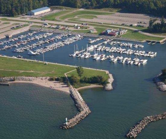 Aerial view of a marina with multiple docks filled with boats, surrounded by parking lots, grassy areas, and a breakwater protecting the harbor from a large body of water. Trees and a few buildings are visible nearby.