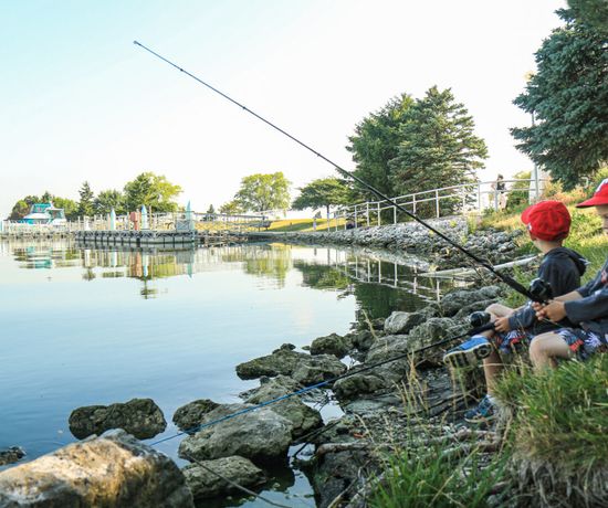 Two young boys sit on rocks by a calm lakeshore, fishing with rods. They wear casual clothes and red caps. Trees, a railing, and a docked pier are visible in the background on a sunny day.
