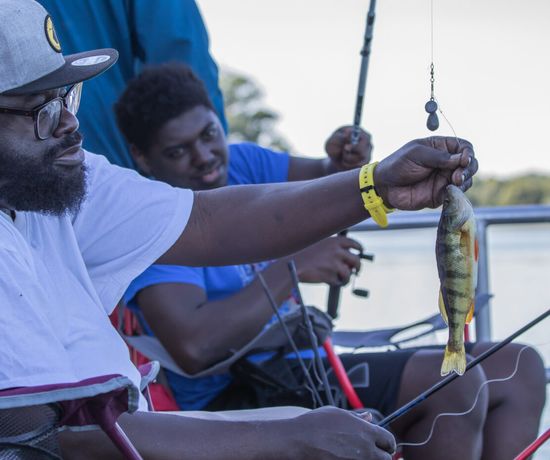 Two men sit on a dock fishing; one man in the foreground holds up a small yellow perch he has caught, while the other man smiles and holds his fishing rod. Trees and water are visible in the background.