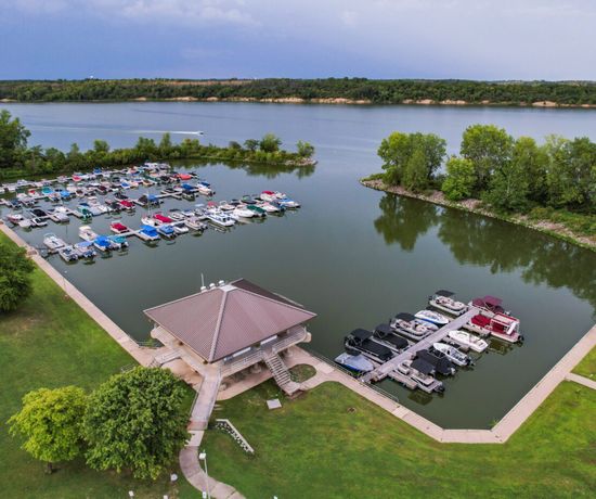 Aerial view of a marina with boats docked at piers, surrounded by green trees and grassy areas next to a large lake under a cloudy sky.