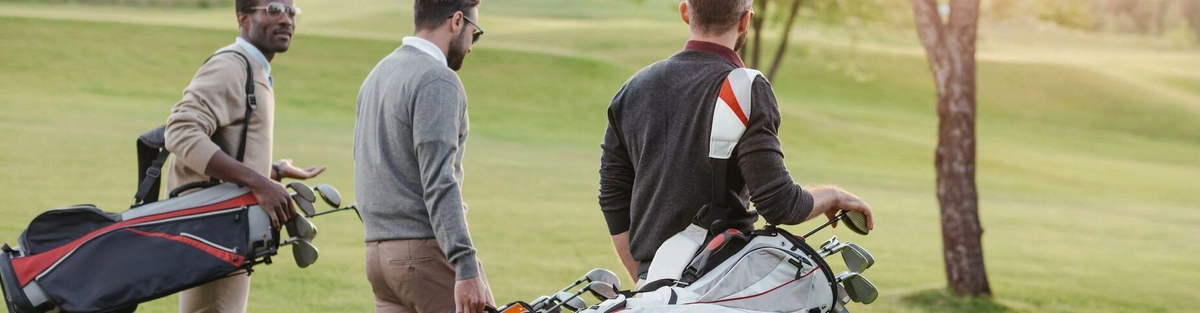 Three men carrying golf bags walk together on a grassy golf course, with trees and sunlight in the background. They appear to be having a conversation while heading towards the next hole.