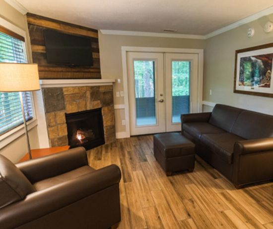 A cozy living room with two brown leather sofas, a matching ottoman, a stone fireplace with a TV above it, wooden floors, a framed wall picture, and glass doors leading outside, surrounded by windows.