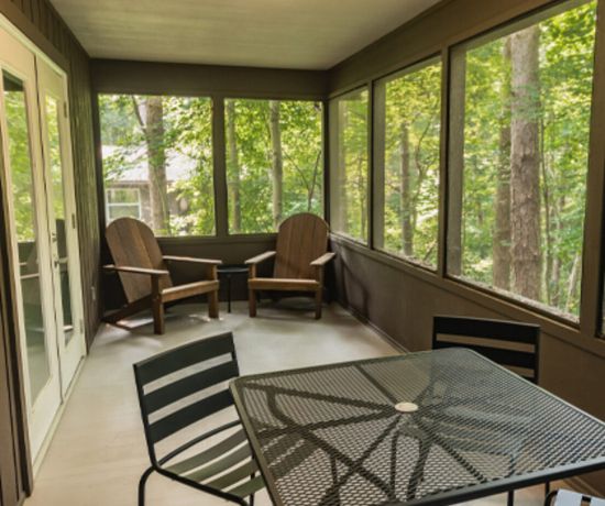A screened-in porch with two wooden Adirondack chairs by the windows and a black metal table with four matching chairs. The porch overlooks a lush, green wooded area.
