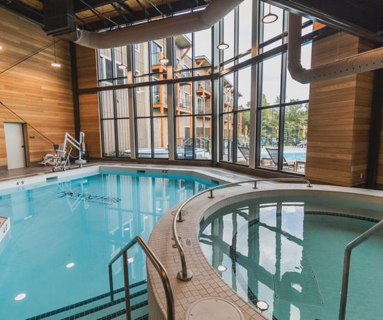 Indoor pool area with large windows, a round hot tub in the foreground, and a rectangular swimming pool behind it. Wooden walls and ceiling create a warm atmosphere; pool chairs and outdoor view are visible through the glass.