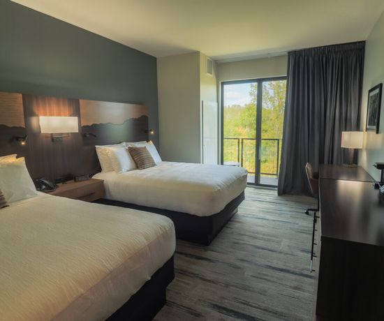 A modern hotel room with two double beds, a desk, lamps, a large wall-mounted TV, and a window with dark curtains overlooking green trees outside.