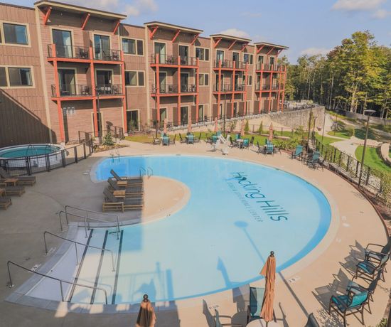 A curved outdoor pool with lounge chairs and umbrellas is surrounded by a modern, three-story apartment building with balconies, adjacent hot tub, and landscaped green areas under a sunny sky.