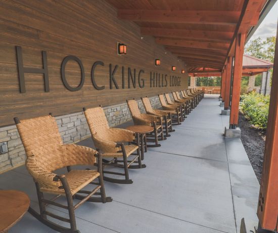 A row of wicker rocking chairs lines a covered outdoor patio at Hocking Hills Lodge, with a stone and wood exterior and small wall lanterns.