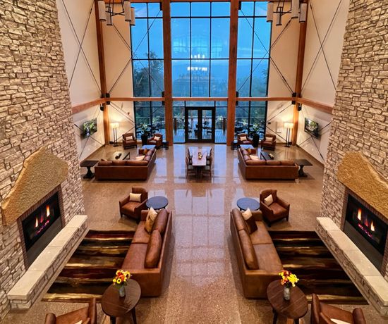 Spacious lodge lobby with high ceilings, stone walls, modern fireplaces, cozy seating areas, and large windows offering a scenic view of trees and a lake outside. Warm, inviting atmosphere with soft lighting and contemporary decor.