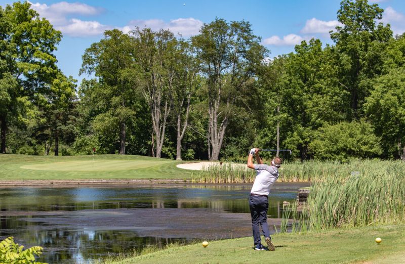 A golfer in mid-swing stands near a pond, facing a green surrounded by trees under a bright blue sky with scattered clouds. The golf course is lush and scenic.