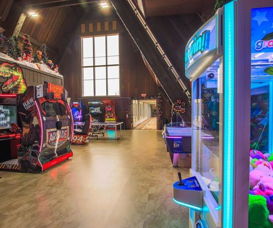 A brightly lit arcade with various gaming machines, including a Jurassic Park game, air hockey table, and a claw machine filled with colorful plush toys. The room has high ceilings and holiday decorations.