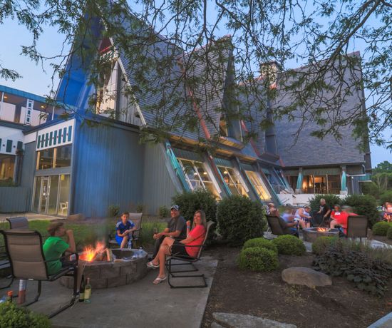 People sit around outdoor fire pits in front of a modern, triangular-roofed building at dusk, relaxing in patio chairs amid greenery, with trees and lights creating a cozy atmosphere.