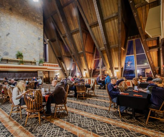 A warmly lit restaurant with high, sloped wooden ceilings and large windows. Diners sit at tables with wooden chairs, engaged in conversation, while soft light creates a cozy, inviting atmosphere.