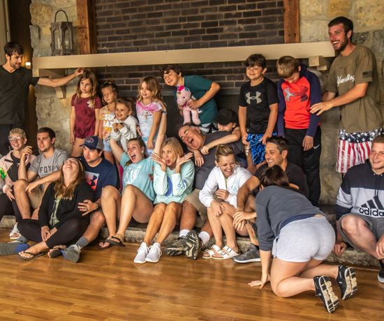 A large group of adults and children sit and kneel together indoors in front of a stone fireplace, laughing and making silly faces, creating a playful and joyful atmosphere.