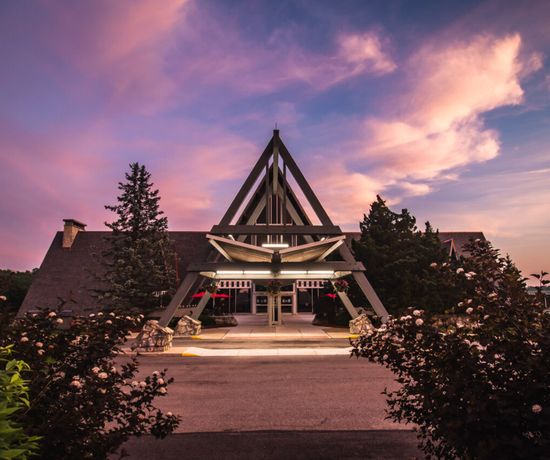A-frame building with a prominent triangular entrance, surrounded by bushes and flowering plants, under a vibrant, colorful sunset sky with pink and purple clouds.