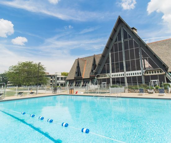 A large outdoor swimming pool with blue water sits in front of a modern building with tall, triangular peaks and many windows under a blue sky with scattered clouds. Lounge chairs and trees are visible in the background.