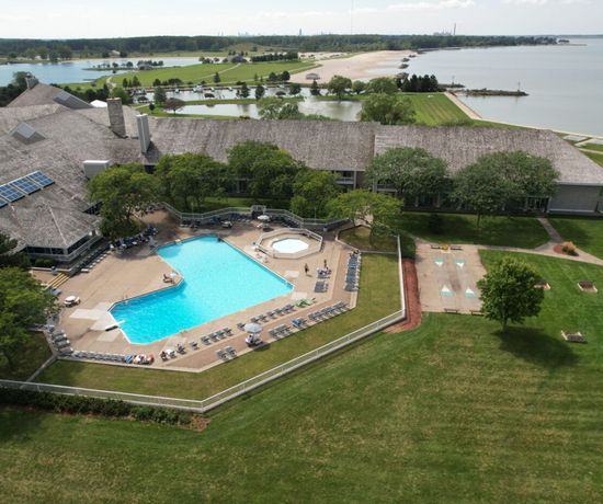 Aerial view of a large lakeside resort featuring an outdoor swimming pool, hot tub, sun loungers, and shuffleboard courts on a grassy lawn, with a sandy beach and water in the background.
