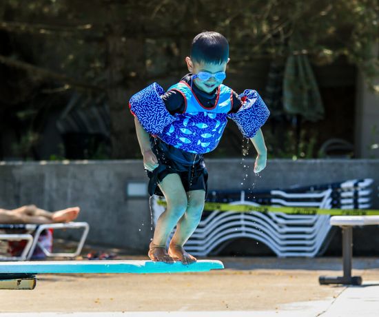 A young child wearing blue floaties and goggles stands on the edge of a diving board, preparing to jump into a pool. Lounge chairs and people relaxing are visible in the background.