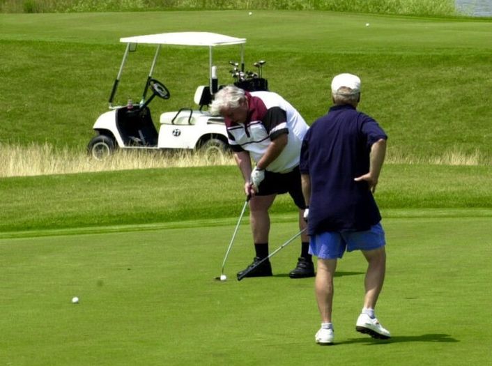 Two men on a golf course; one is putting while the other watches. A golf cart and golf clubs are visible in the background on the grass.