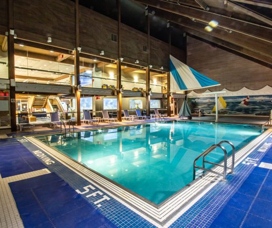 Indoor swimming pool with clear water, surrounded by blue non-slip mats. Lounge chairs line the sides, and large windows offer views of a modern, well-lit interior with wood accents and nautical decor.
