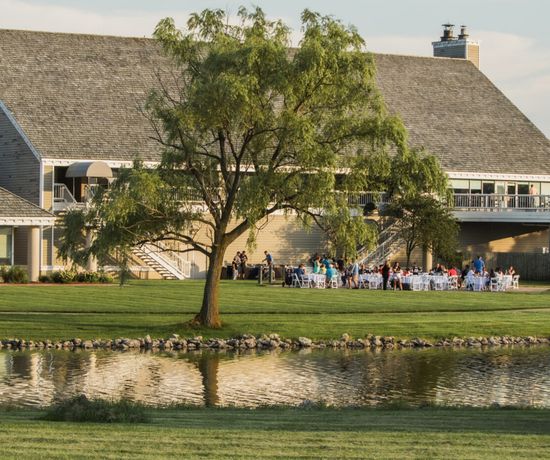 A group of people gather around white chairs and tables on a lawn near a pond, outside a large building with a balcony and a tree in the foreground, on a sunny day.