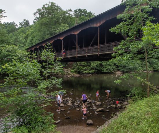Children and adults play on rocks in a shallow creek beneath a large, wooden covered bridge surrounded by lush green trees.