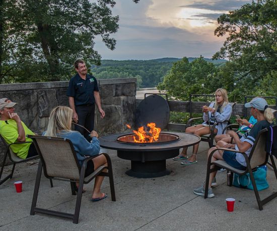 Five people sit around a fire pit outdoors, chatting and relaxing on patio chairs with drinks. Trees and a scenic, hilly landscape are visible in the background at sunset.