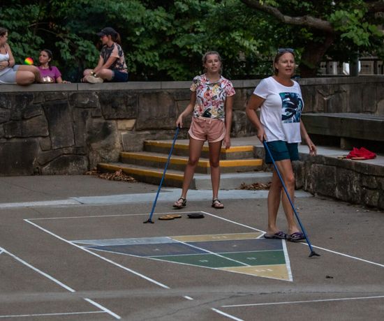 Two women play shuffleboard on an outdoor court while three people sit and talk on stone steps nearby, surrounded by trees and a stone wall.