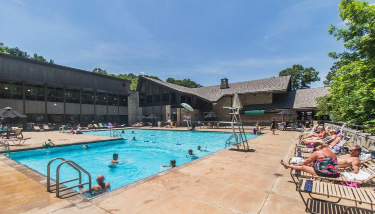 Outdoor swimming pool surrounded by people lounging on chairs and swimming, next to a large lodge building with stone and wood exterior, under a clear blue sky and bright sunlight.