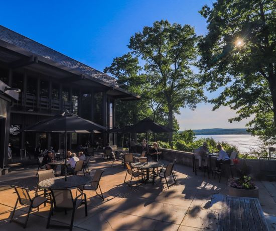 People sit at tables on a sunny outdoor patio with umbrellas, surrounded by trees and overlooking a lake. Sunlight filters through the leaves, creating a relaxed, scenic atmosphere.