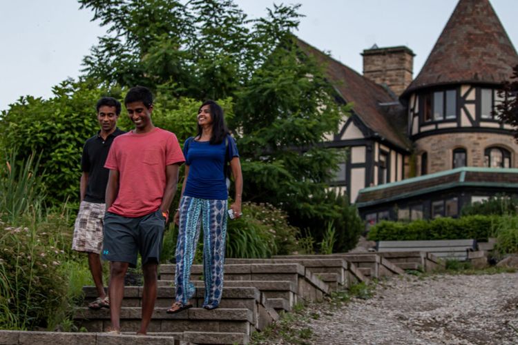 Three people walk down outdoor stone steps, smiling and chatting, with lush greenery and a large, old stone house with a turret and timber accents in the background.