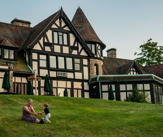 An older woman and a young child hold hands while sitting and standing on a grassy hill in front of a large, Tudor-style house with dark wooden beams and steep roofs.
