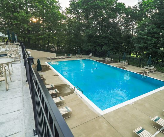 A rectangular outdoor swimming pool surrounded by lounge chairs and umbrellas, next to a patio with tables and chairs, bordered by trees and greenery. The pool area is empty and appears clean and well-maintained.