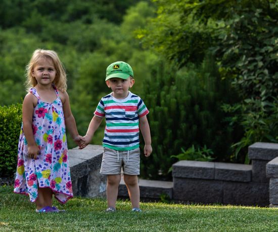 Two young children stand on grass, holding hands. The girl wears a colorful floral dress and purple sandals; the boy wears a green cap, striped shirt, and shorts. They are outdoors with greenery and stone steps behind them.